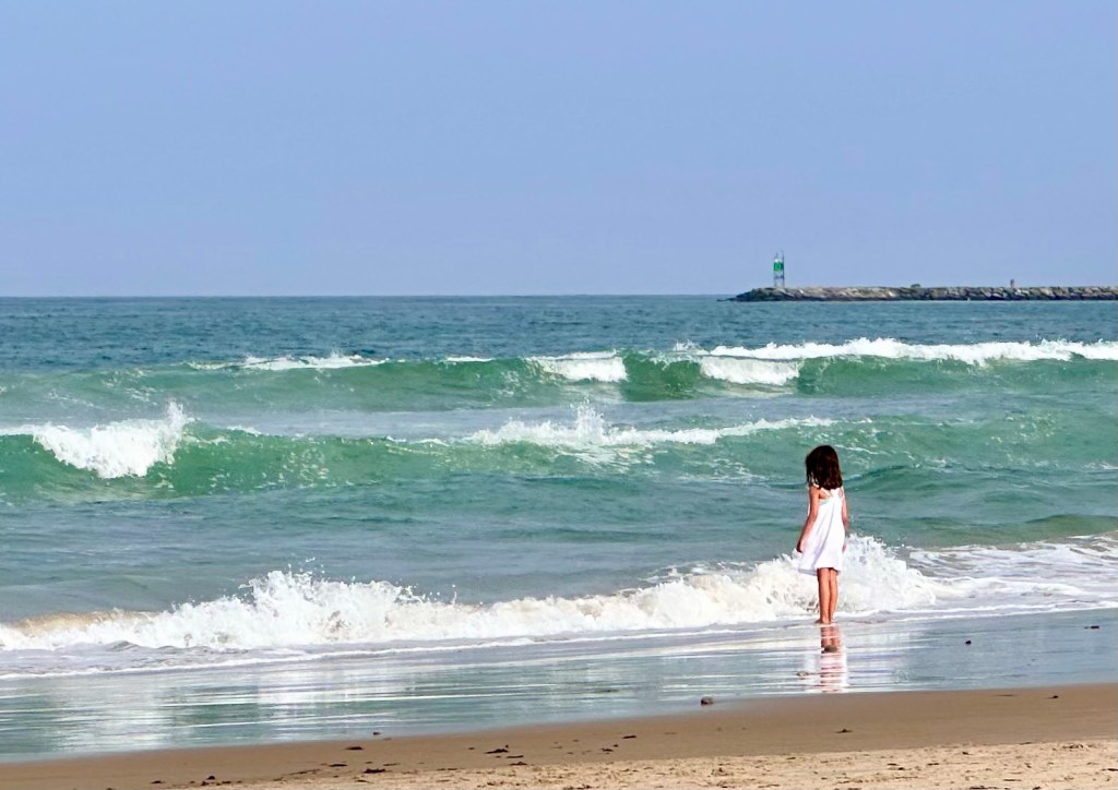 A young girl in a white dress standing at the edge of the ocean, facing the rolling waves with quiet bravery.