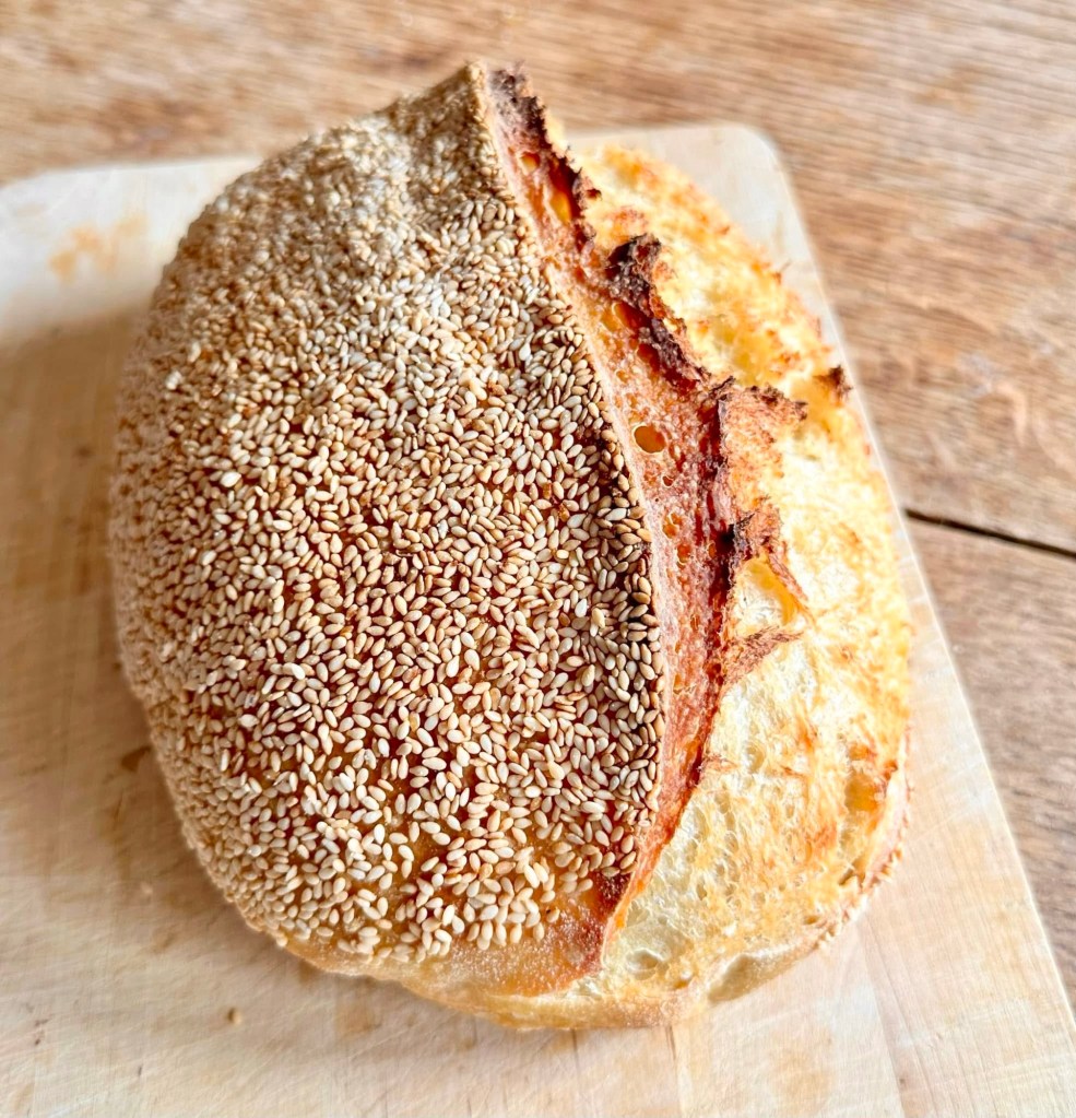 Sourdough batard topped with sesame seeds on wooden cutting board.