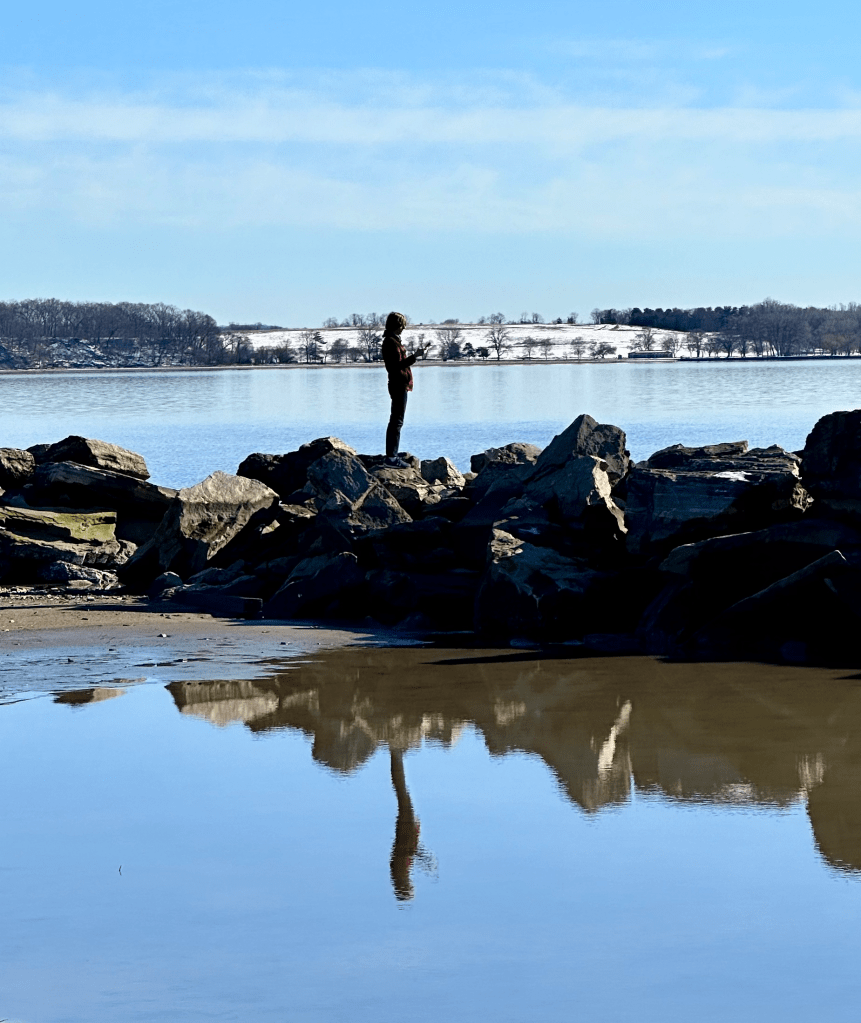 Silhouetted figure standing on rocks in the Hudson River with still water reflecting the sky.