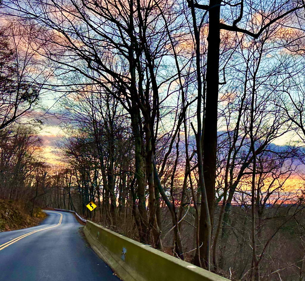 Curving road through wooded landscape with silhouetted trees and a colorful sunset sky.