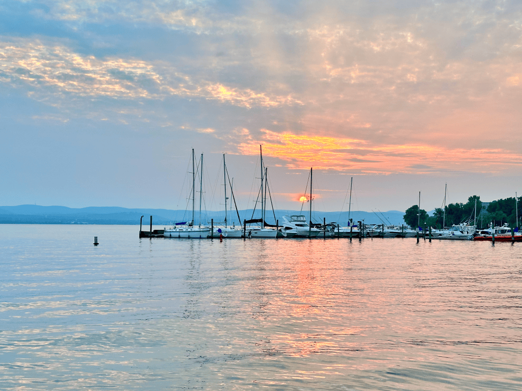 A serene lakeside view featuring multiple sailboats docked along a pier during sunset, with a colorful sky reflecting in the water.