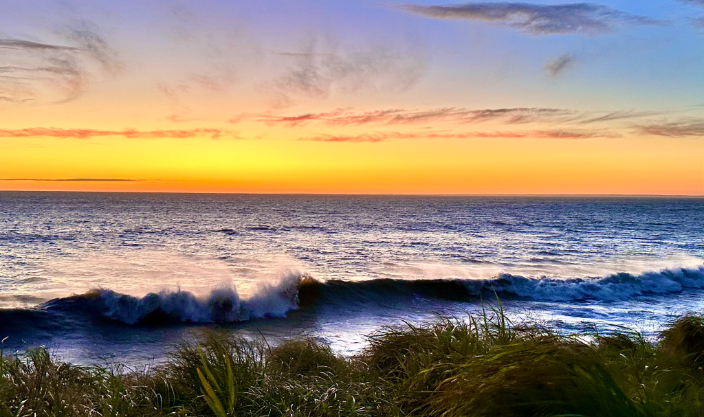 Golden sunset over the ocean waves breaking on the shore and tall dune grass in the foreground.