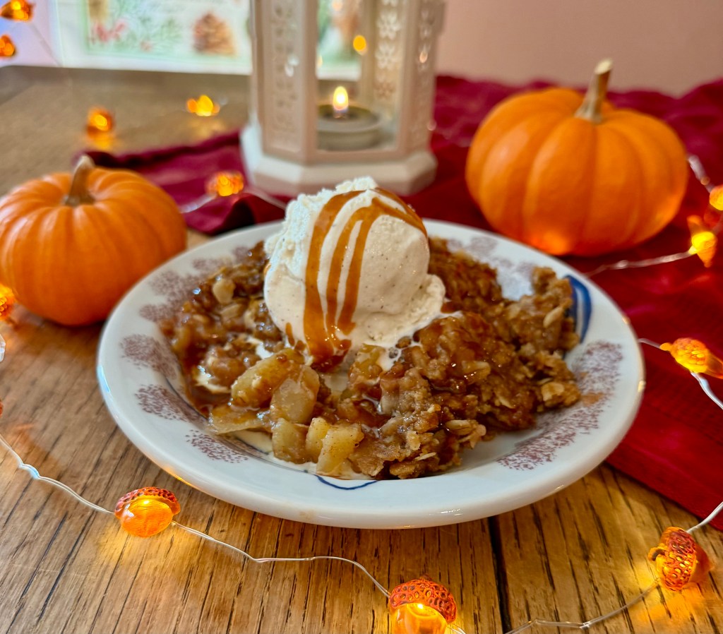 Warm apple crisp topped with a scoop of vanilla ice cream and drizzled salted caramel, served in a cozy fall setting with mini pumpkins and soft candlelight.