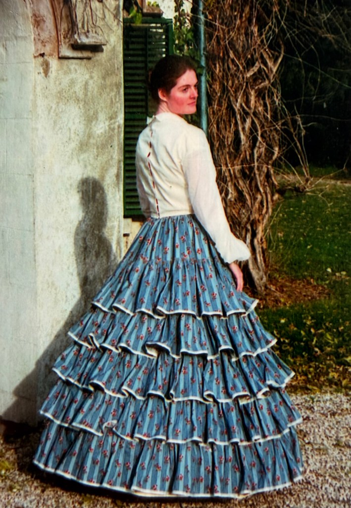 A young woman wearing a blue and white dress in the vVctorian style of the 1850's - standing in front of Sunnyside cottage in Tarrytown, NY.