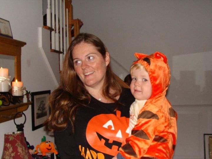A woman in a Halloween-themed shirt holds a child dressed in an orange tiger costume, standing in a home decorated for Halloween.