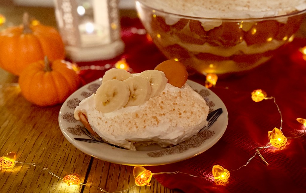 A serving of banana pudding displayed in a warm setting of lights, candle, pumpkins and layered banana pudding trifle in background.