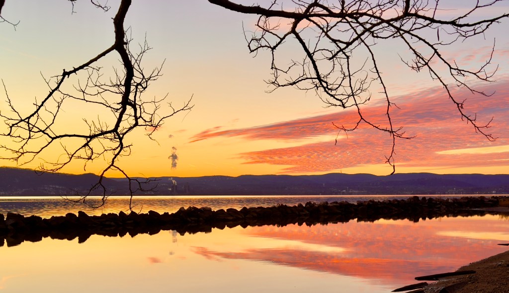 A pink and gold sunset reflecting on a glassy Hudson River in winter - all framed by the bare branches of tree.