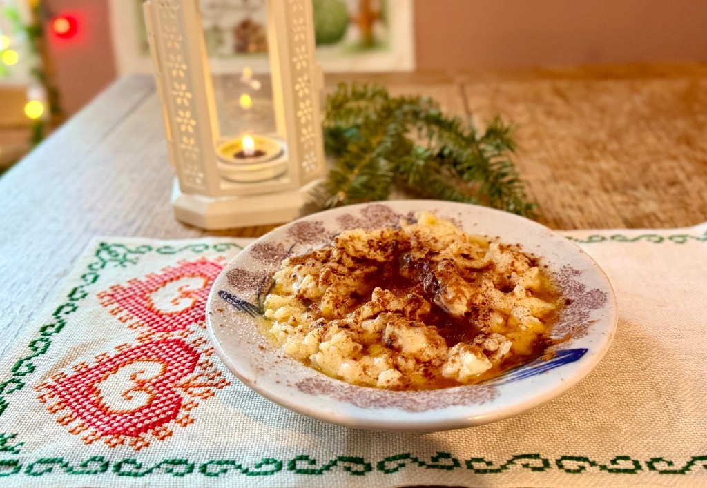 Traditional Norwegian creamy rice pudding - Risgrøt - flavored with cardamom and vanilla then topped with melted butter, sugar and cinnamon. Served on blue and white china with a traditional Norwegian cross stitch table runner on a rustic farmhouse table.