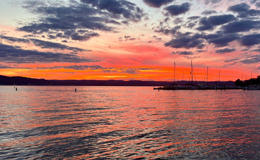 Dramatic sunset over a tranquil body of water, with vibrant orange and pink hues in the sky, and silhouettes of sailboats in the foreground.