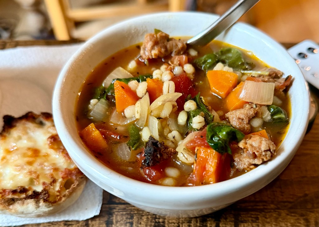 A bowl of hearty vegetable and barley soup featuring chunks of meat, carrots, greens, and tomatoes, accompanied by a slice of cheesy bread on a napkin.