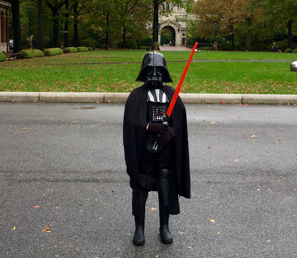 A child dressed as a character from a popular science fiction franchise, wearing a black cape and helmet, holding a red lightsaber, standing in a park with green grass and trees in the background.