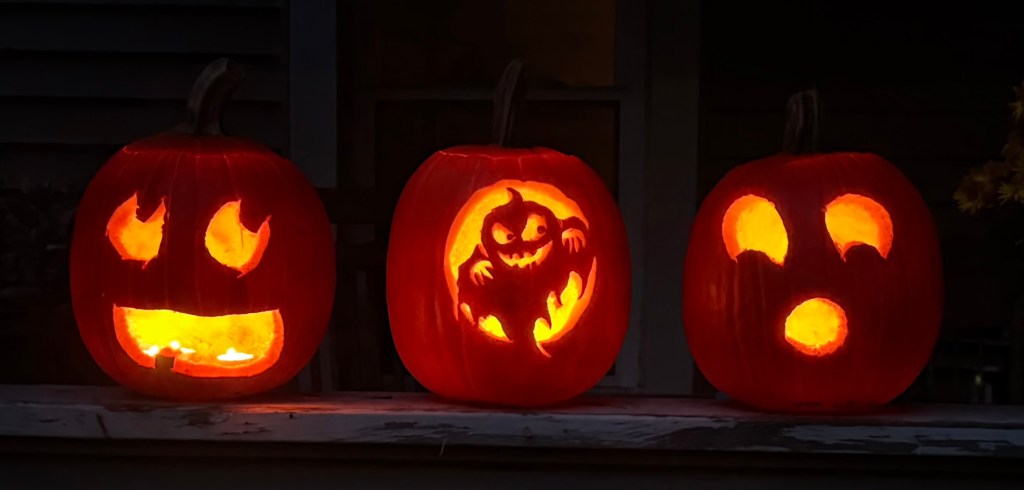 Three carved pumpkins on a porch, glowing with orange light. The left pumpkin has a smiling face, the middle pumpkin features a ghost design, and the right pumpkin displays a surprised expression.