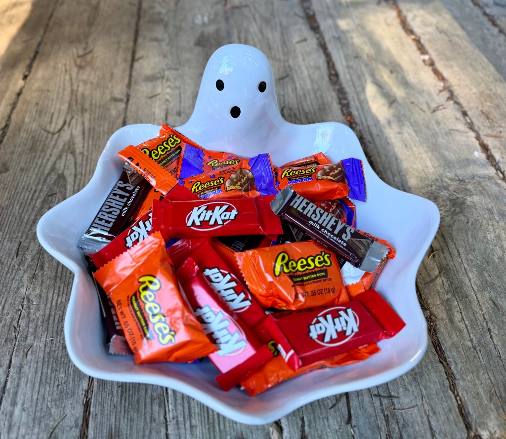 A white ghost-shaped bowl filled with assorted Halloween candies including Reese's, Kit Kat, and Hershey's, placed on a wooden surface.