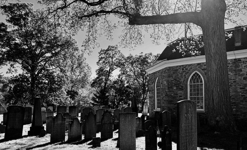 Black and white image of a cemetery with gravestones, a large tree, and a stone church in the background, under clear skies.