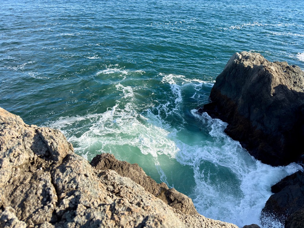 Aerial view of rocky shoreline with waves crashing against the rocks and deep blue ocean water.